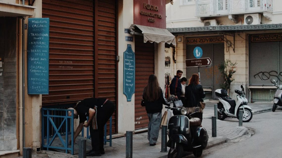 A lively Athens street captures pedestrians, scooters, and shops in the evening light.